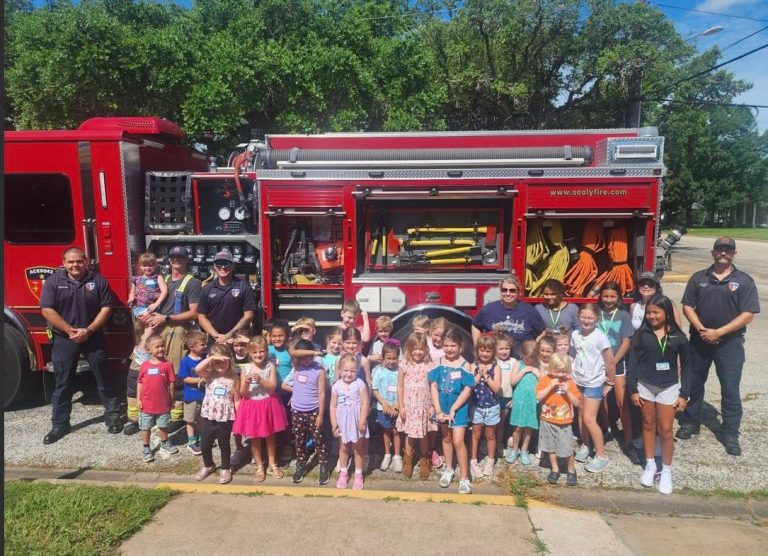 A group of children and four firefighters pose in front of a red fire truck on a sunny day. The kids are smiling, some waving, with trees and a street in the background.